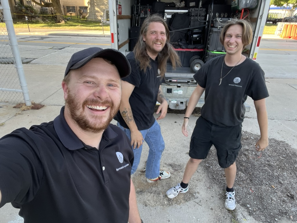 Three Harmonic Productions crew members smiling together beside the equipment trailer