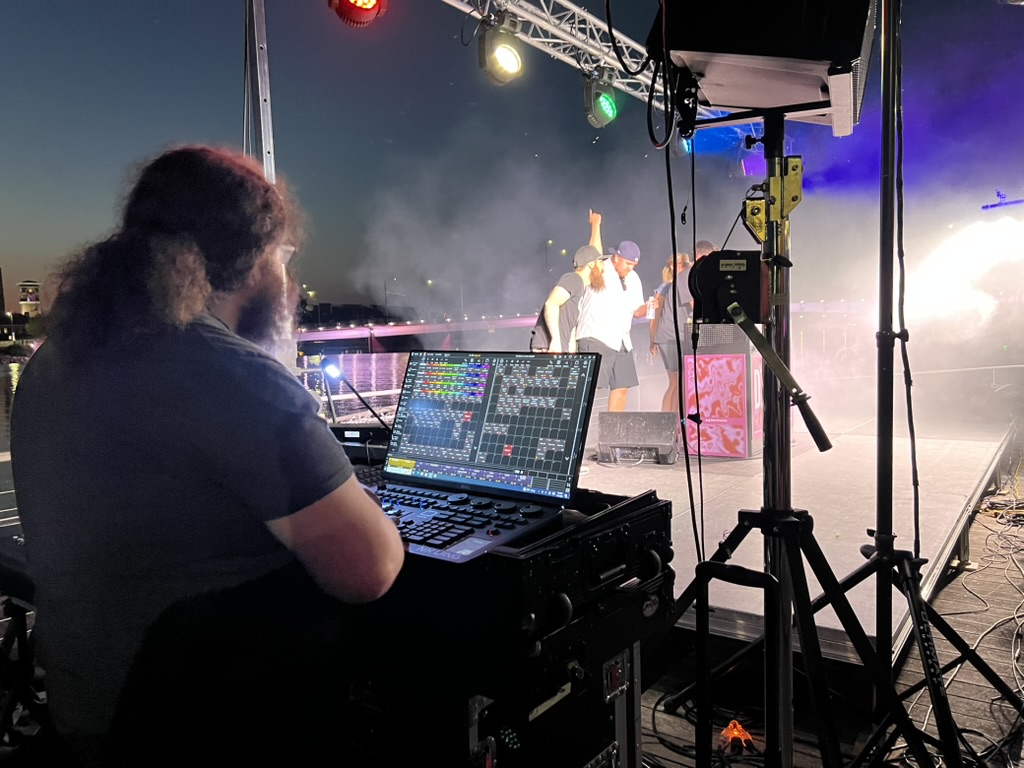 A Harmonic Productions lighting technician operating a console beside the stage