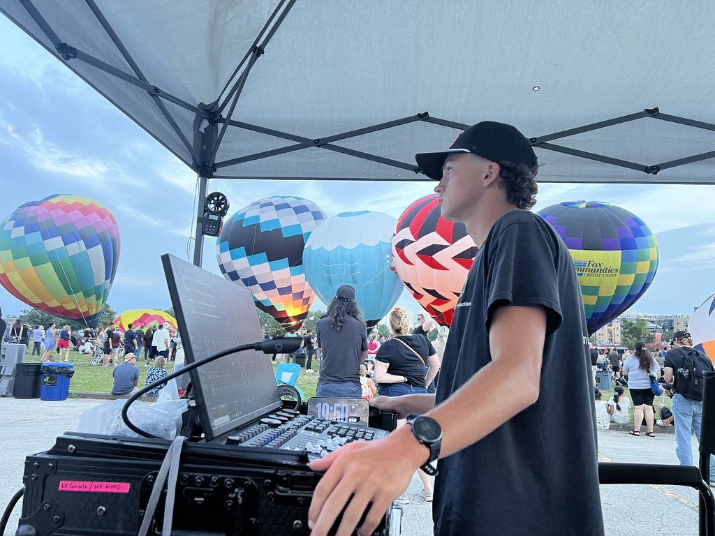 A Harmonic Productions crew member operating event controls with hot air balloons beyond the stage